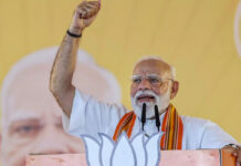 “Why Extra Parliament Session?” Modi Reveals Big Plan for Women Quota & South Seats Prime Minister Narendra Modi standing at a podium decorated with the BJP lotus symbol, raising his right fist in a gesture of victory or determination. He is wearing his signature white half-sleeve kurta and an orange-and-yellow patterned stole (gamcha) during an election campaign rally in April 2026