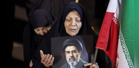 A woman dressed in a black hijab holds a portrait of a bearded cleric wearing glasses and a black turban, alongside an Iranian flag, while appearing to mourn or pray at a public gathering.